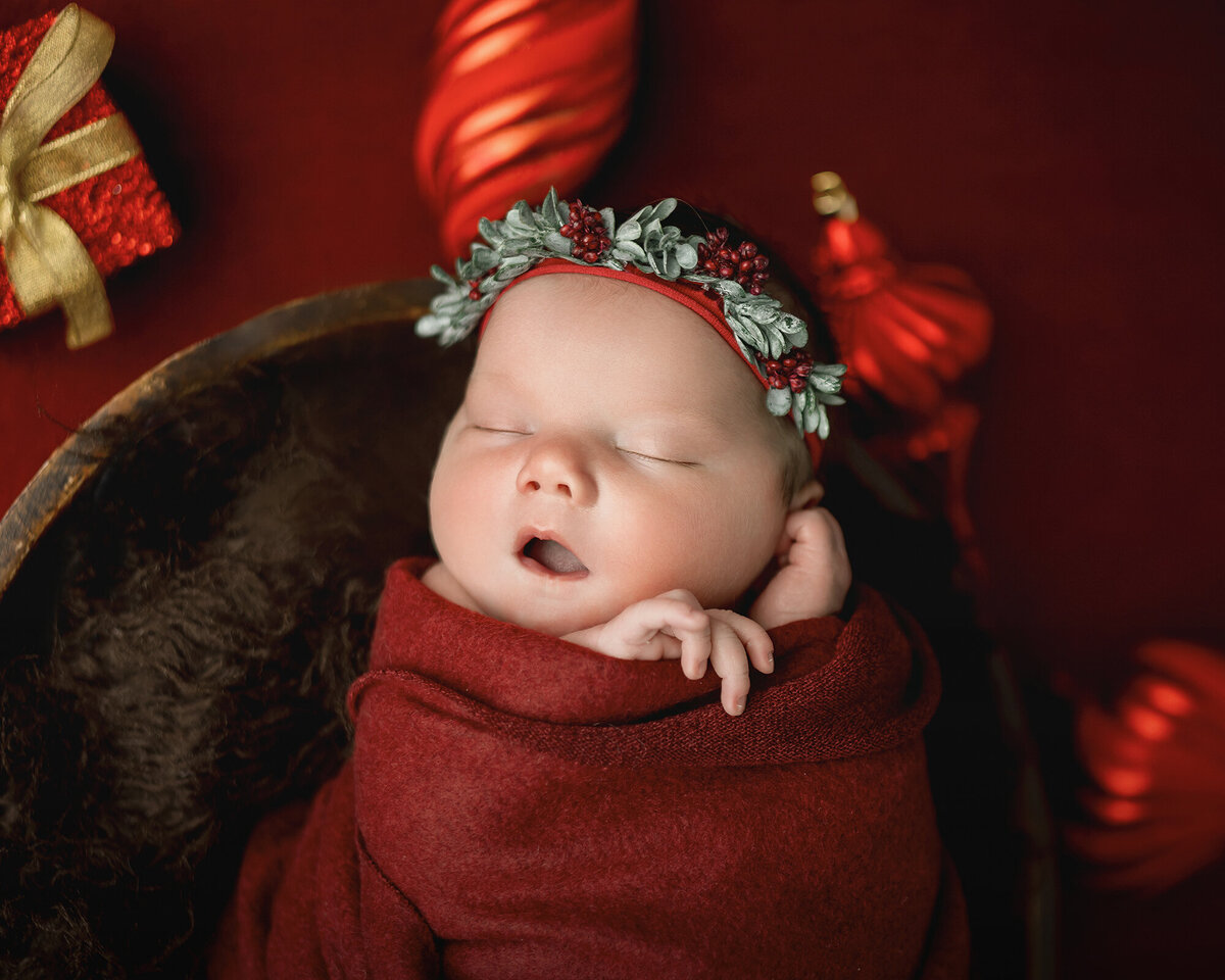 A newborn sleeps with mouth open in a bucket with christmas headband and swaddle
