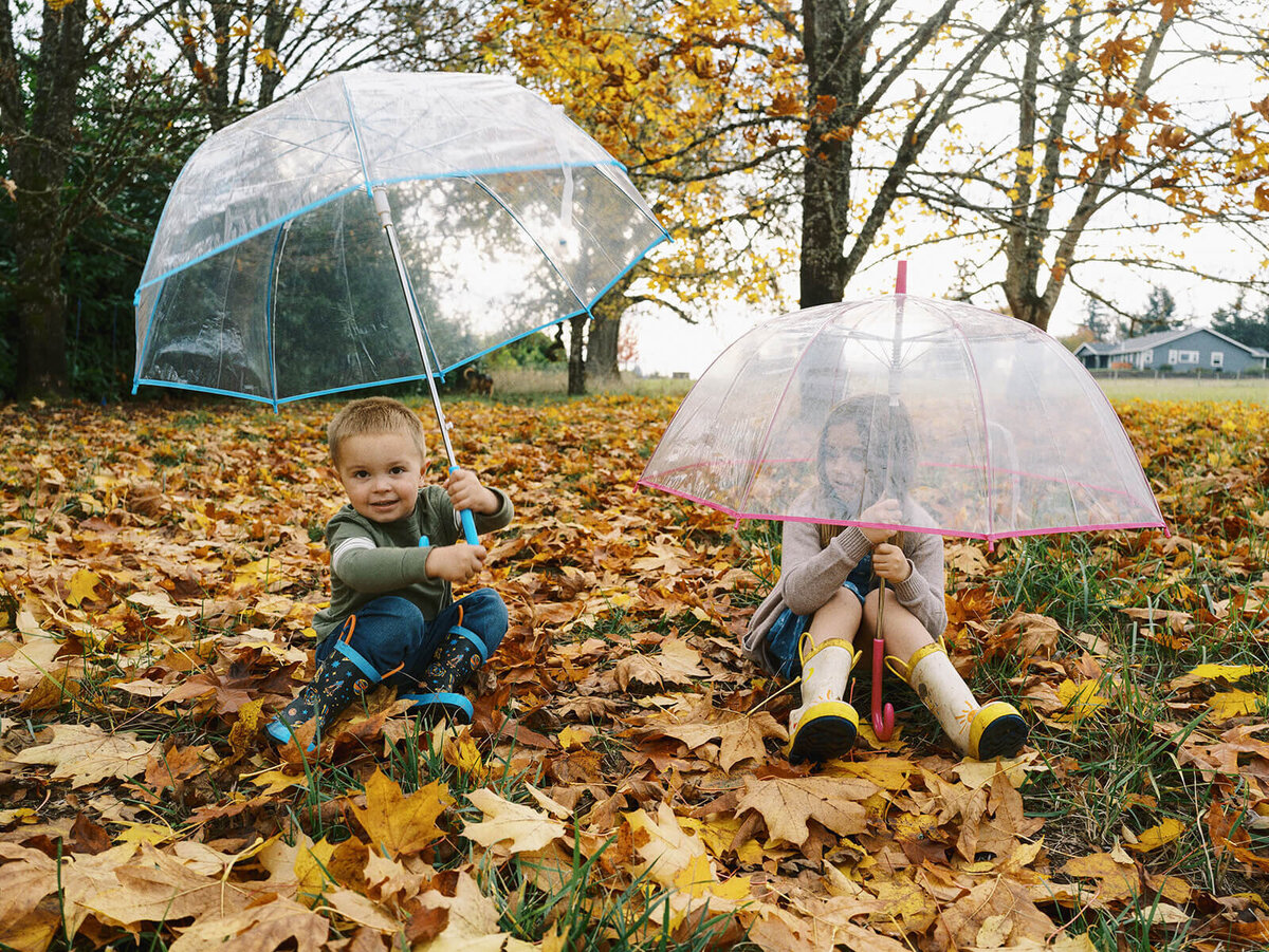 A boy and girl sit outdoor on leaves while holding umbrellas and smiling.