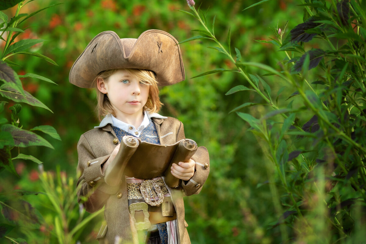 6 year old boy dressed in a pirate costume holding a pirate map.  He is in a jungle-like setting.  He is wearing a tricorn hat and all brown.  He has blond shaggy hair.