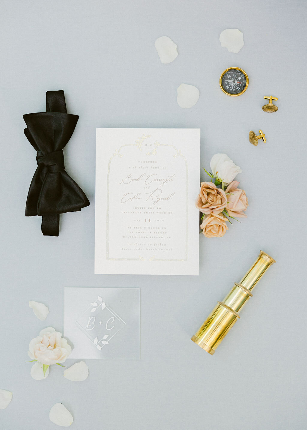 Elegant Bluffton wedding detail flat-lay: ivory invitation, black bow tie, gold telescope, cufflinks, and blush spray roses—captured by luxury wedding photographer Amia Marcell.