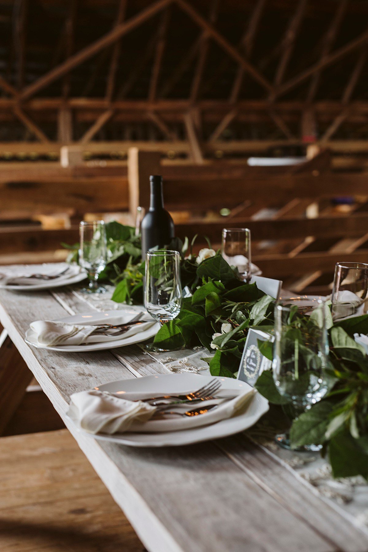 gray barn wood table set with white plates, white linen napkins and gold flatware with a long garlands of greenery, clear crystal water goblets and bottles of wine inside a barn at Arrington Vineyards.