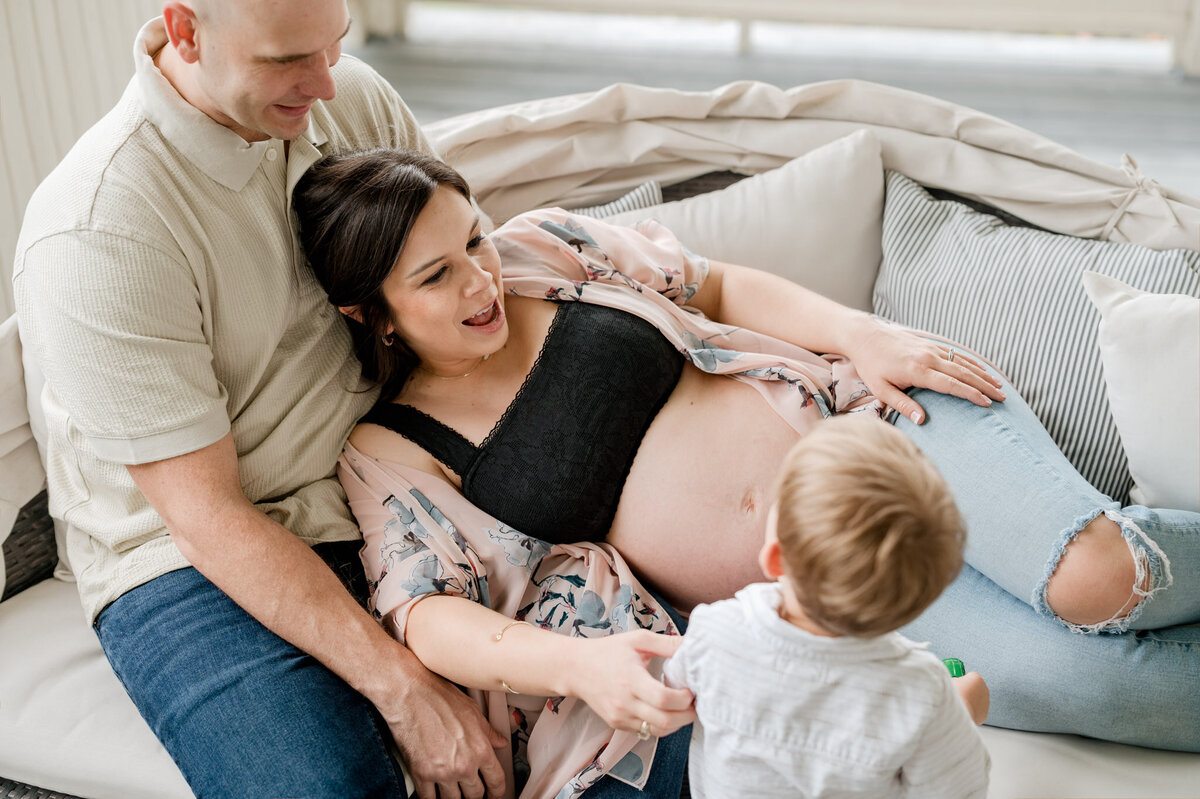 Dad sits on a couch, mom lays against him in a bra and kimono. Their toddler is looking at her growing belly and they smile at him.
