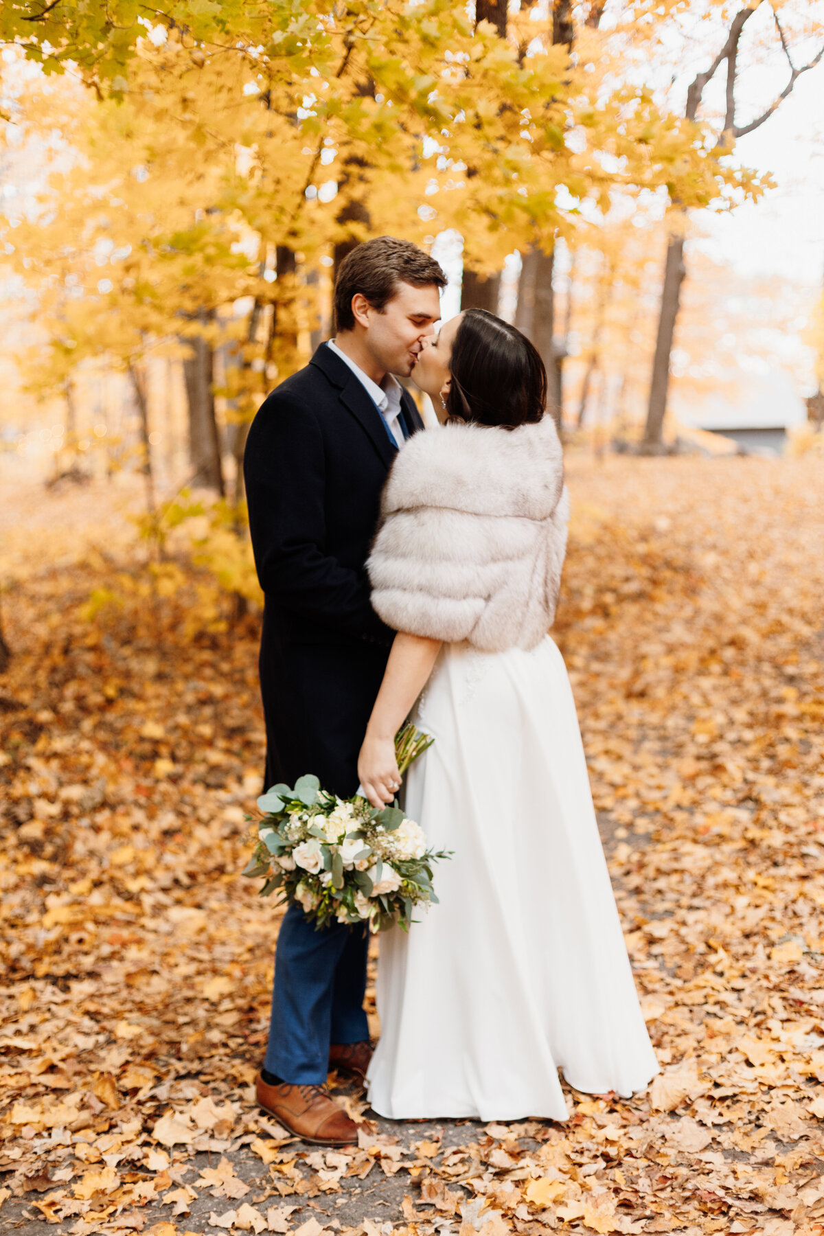 Bride wearing a fur shawl kissing groom underneath a tree with yellow leaves