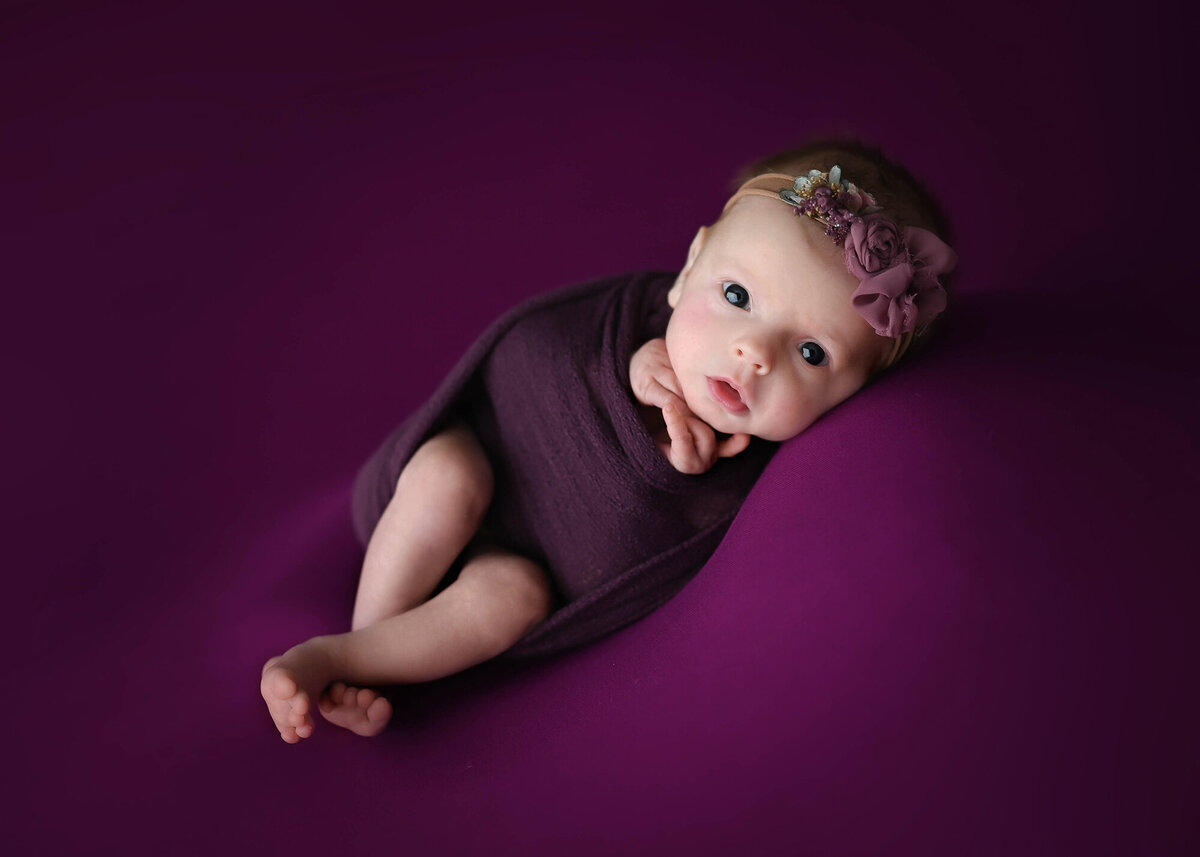 A baby girl in a purple swaddle lays on a matching bed in a studio with a matching bow headband