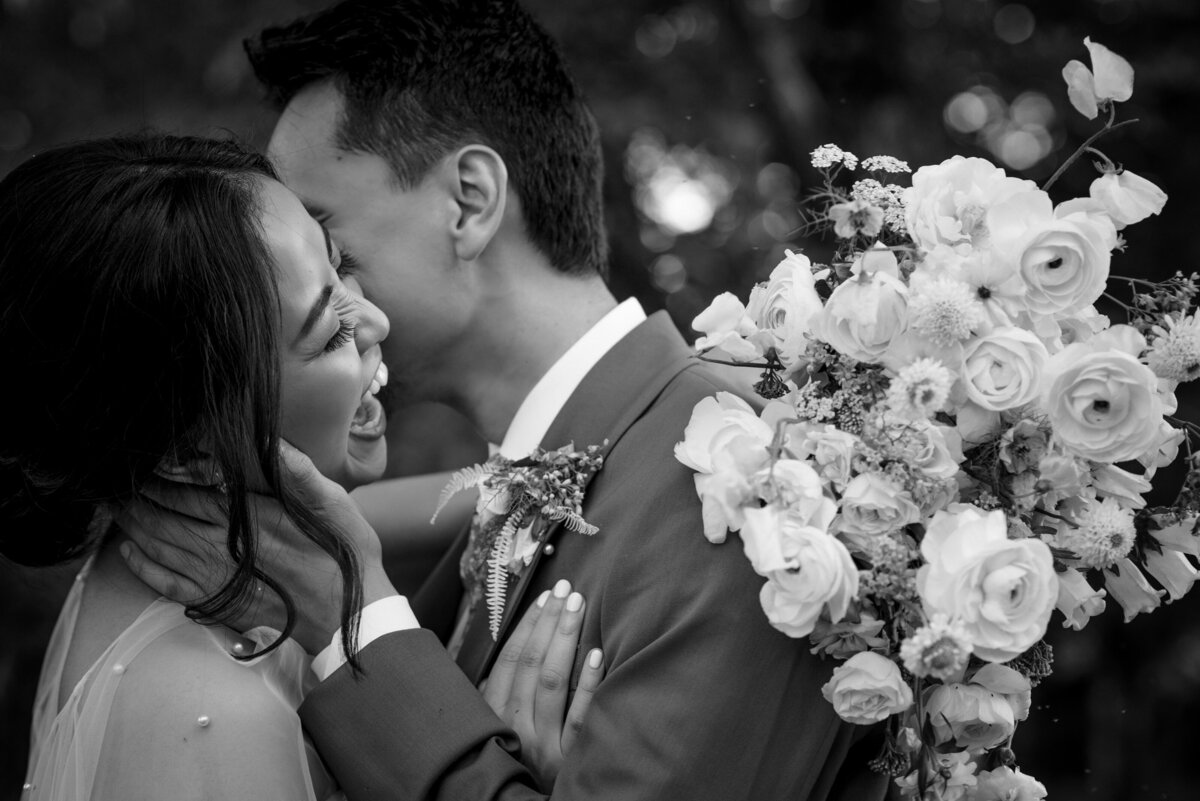 Black and white image of a laughing Asian bride being kissed by groom at The Providence Cotton Mill by Charlotte wedding photographers DeLong Photography
