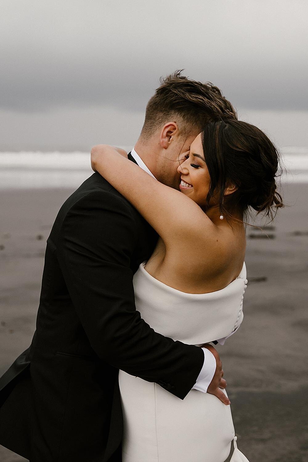 A bride and groom hugging on the opunake beach while aimee kelly photography photographs them