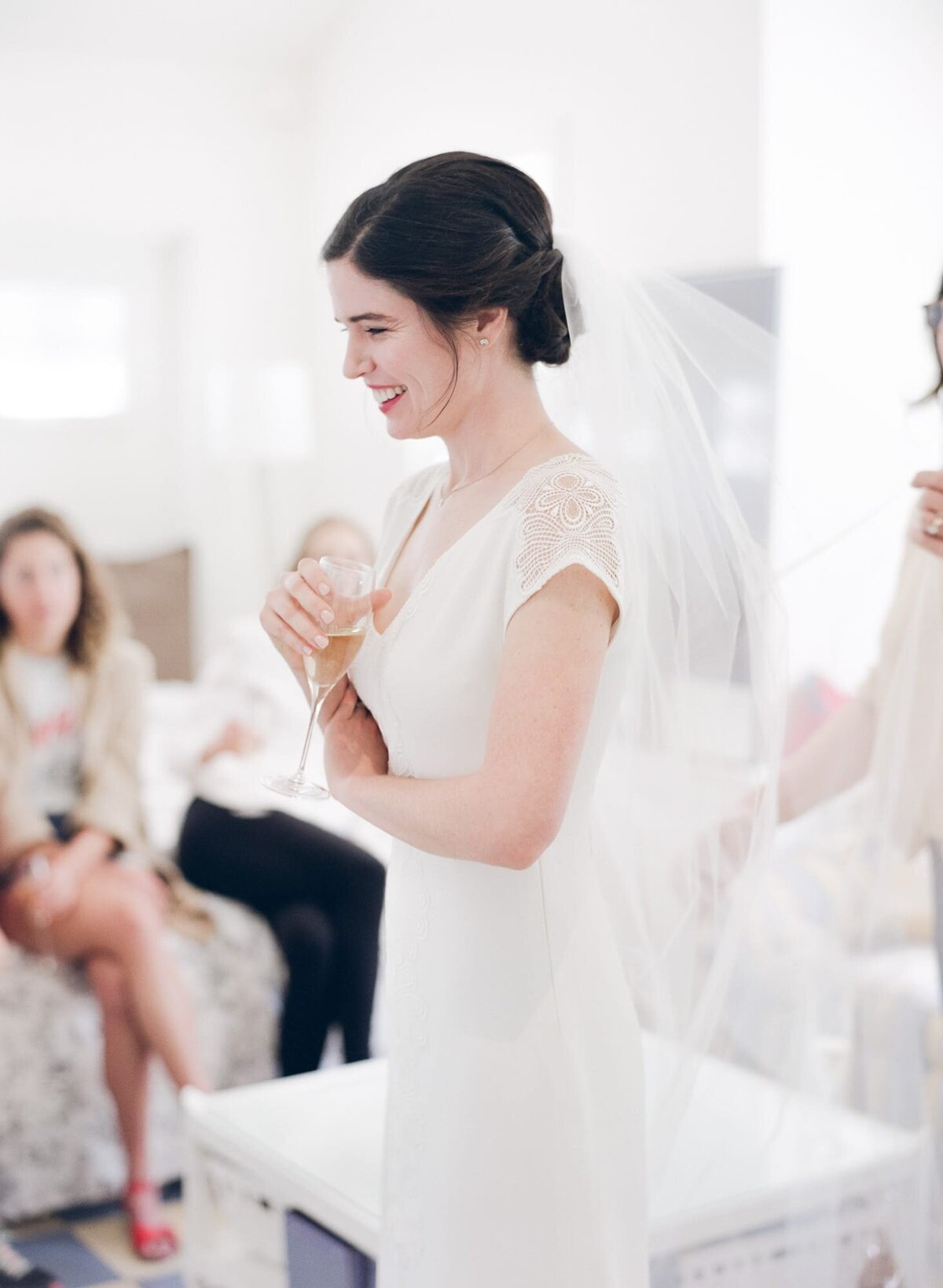 Bride holds a drink in her hand and smiles in anticipation as her maid adjusts her wedding veil.