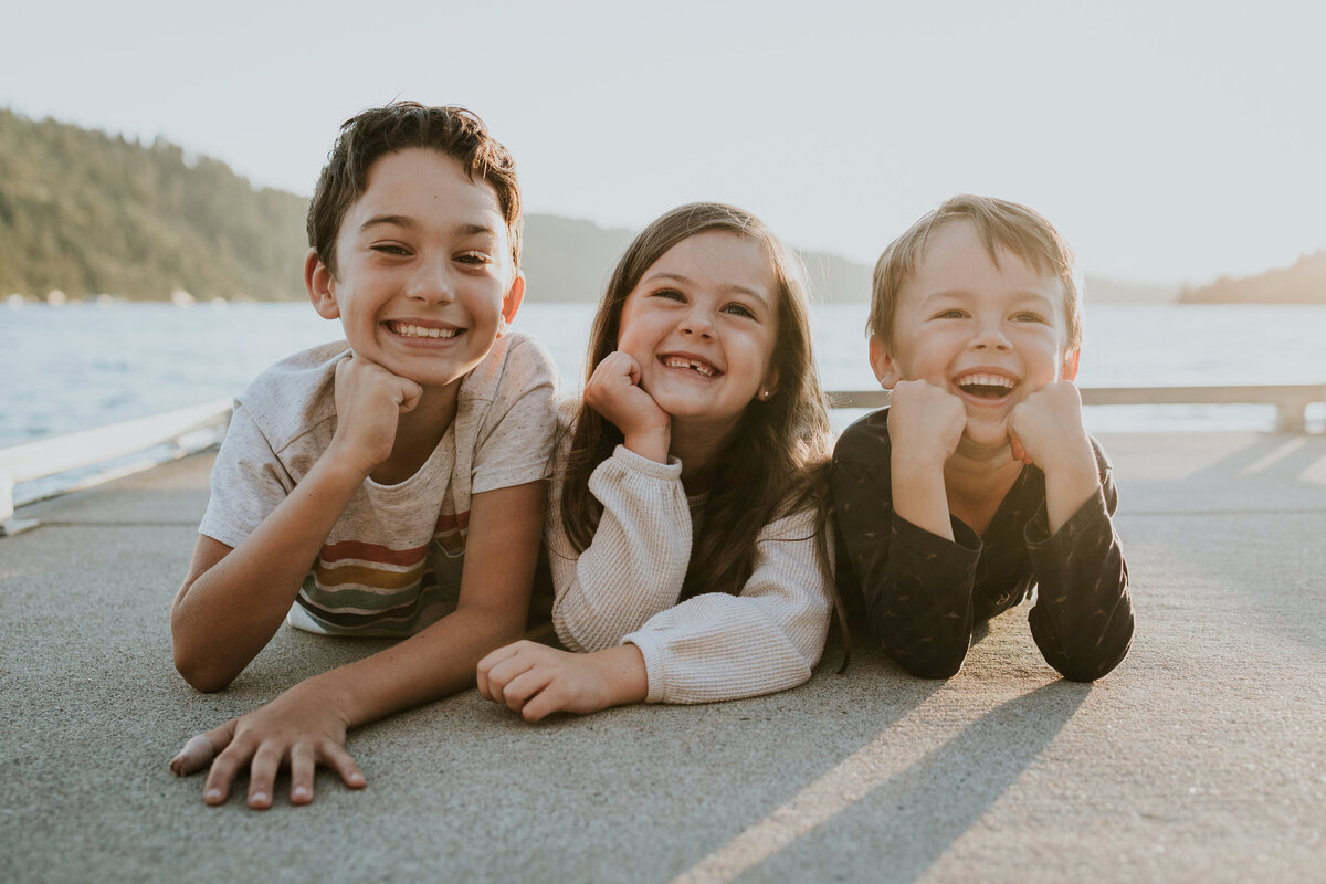 Three young siblings lie on their stomachs together while smiling at camera