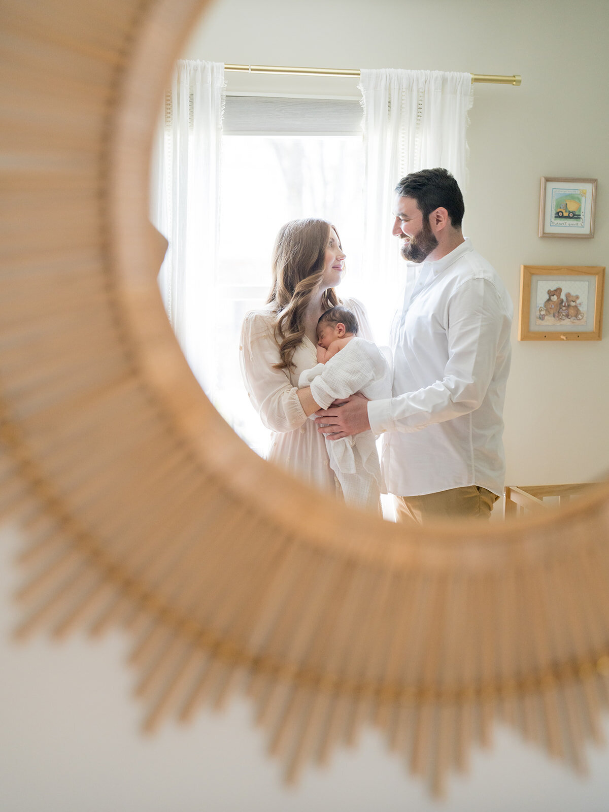 Mother and father stand in the nursery holding newborn baby with a view from the sun mirror.