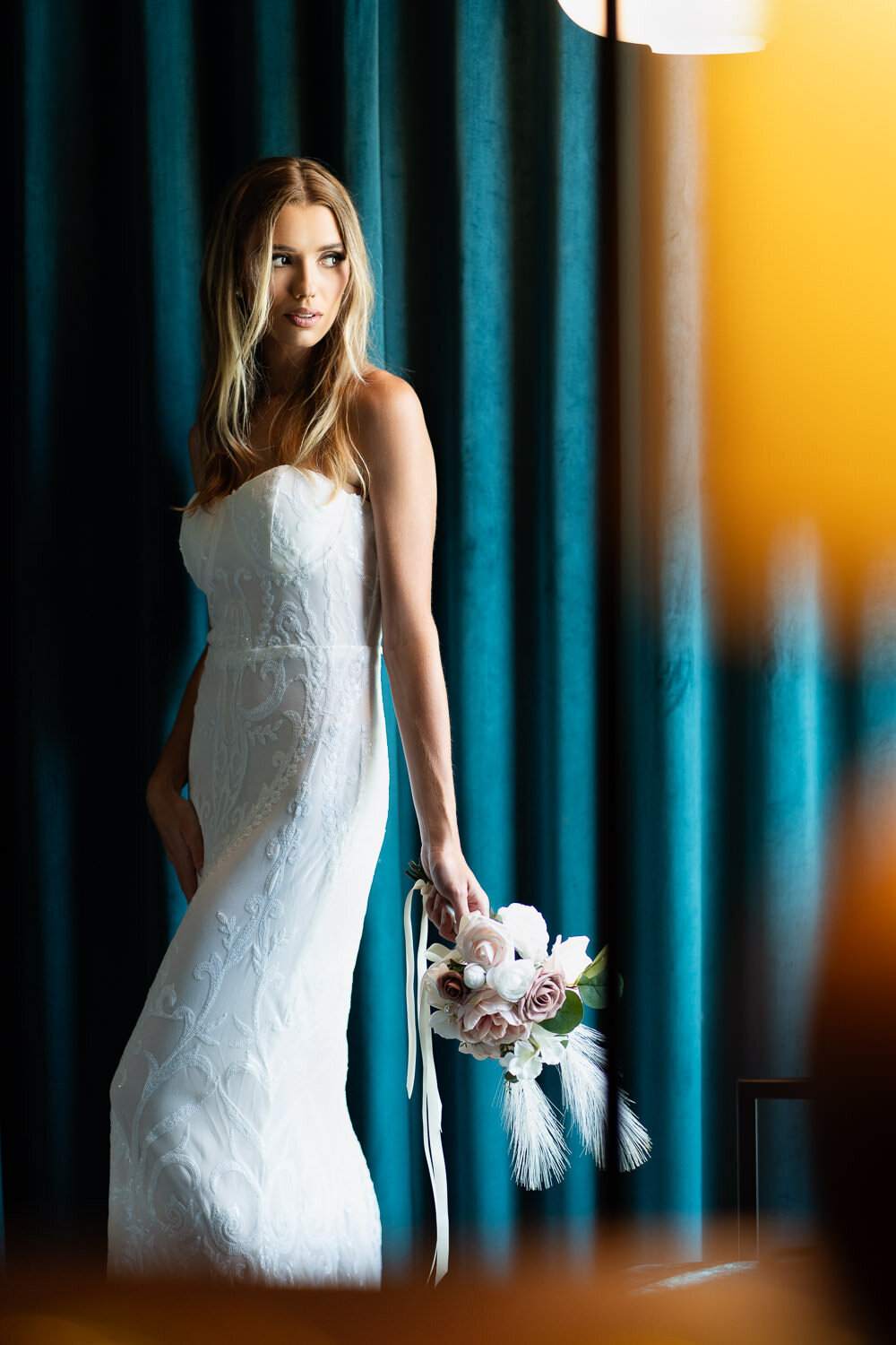 A bride stands in front of rich teal curtains at The Ruby Hotel, softly illuminated by the warm glow of a lamp while holding her bouquet, captured by Art of You Boudoir.