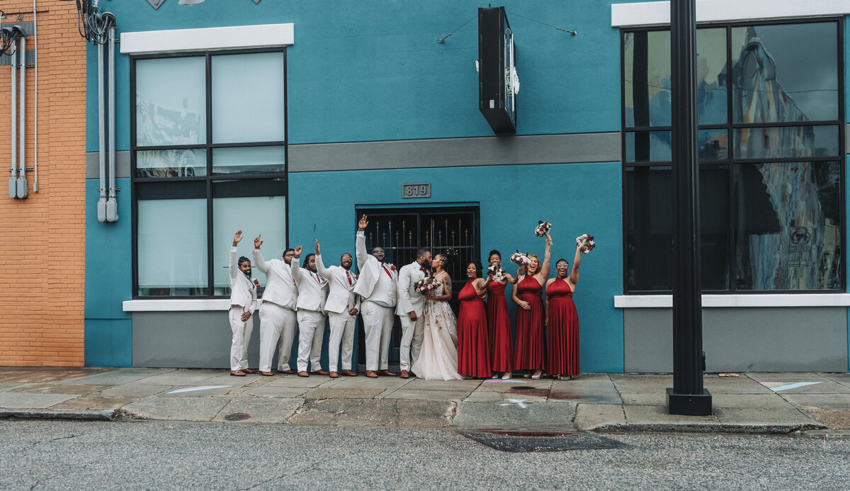A lively and diverse wedding party, with bridesmaids in red dresses and groomsmen in light suits, poses excitedly on the sidewalk in front of the vibrant blue building of The Neon House in Norfolk, Virginia, captured by Quinn Photography LLC.





