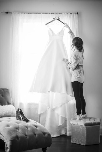 A bride adjusts her wedding gown as it hangs in the window.