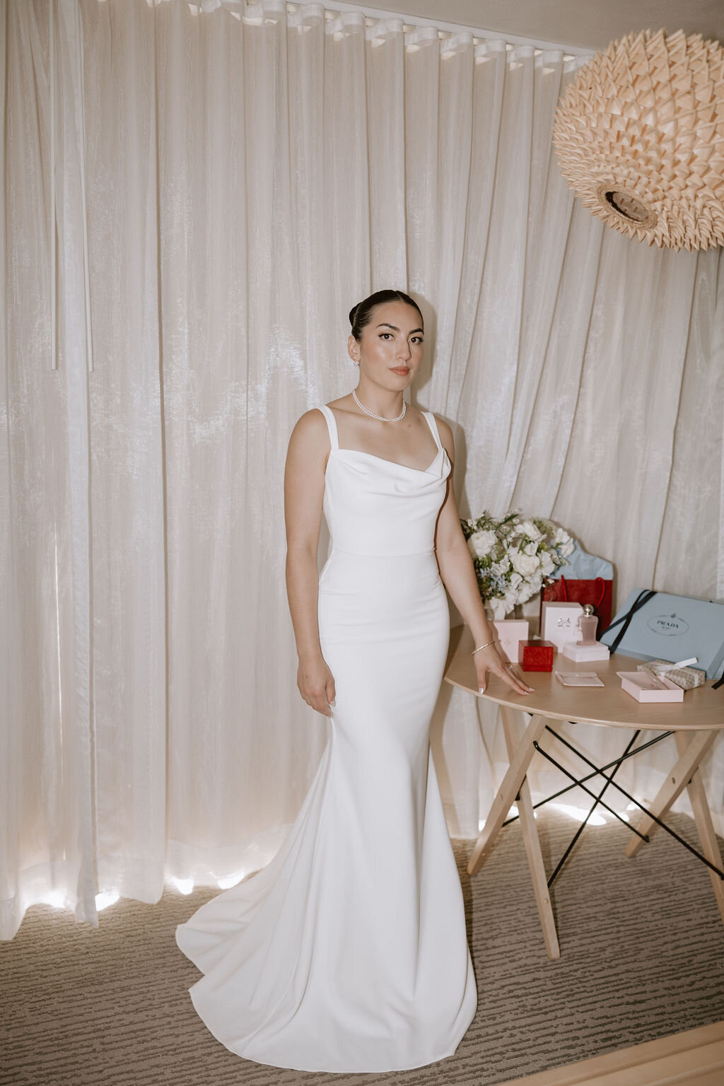 bride in modern grace loves lace wedding dress standing inf ront of curtains and a wooden table in hotel room