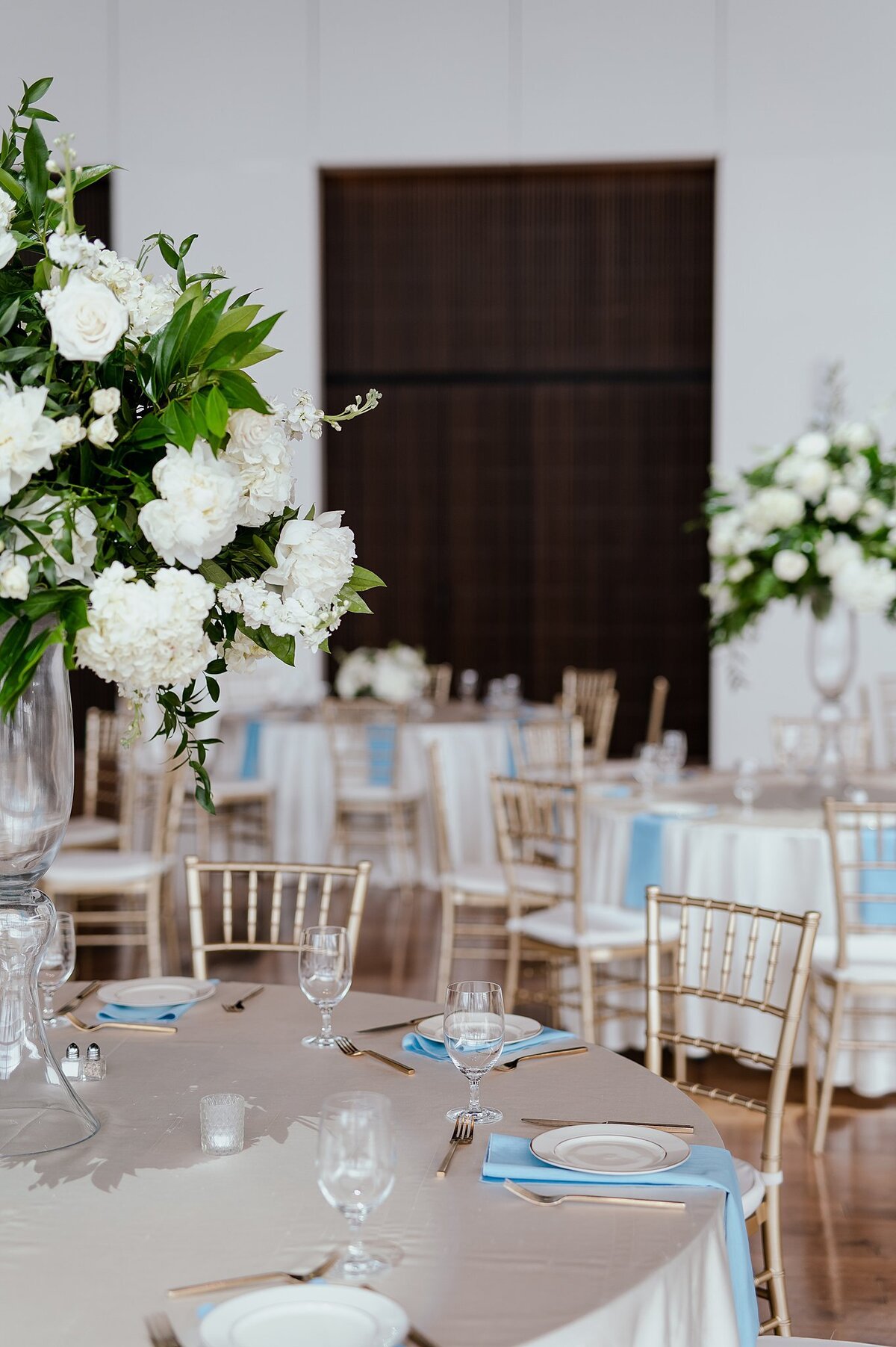 Elegant ivory, blue and gold tables set for a wedding reception at the Country Music Hall of Fame with tall glass vases topped with lush white flowers, greenery and hydrangeas.