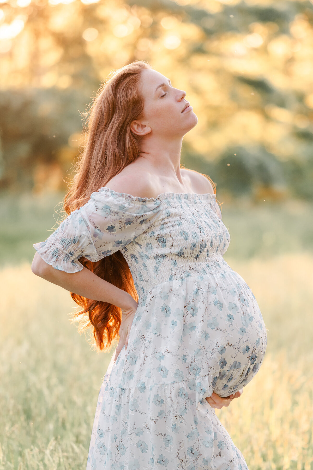 An expectant mother, wearing a white dress with blue flowers, arches her back and tips her head back. She is standing in a grassy field. The setting sun is glowing through the trees behind her.
