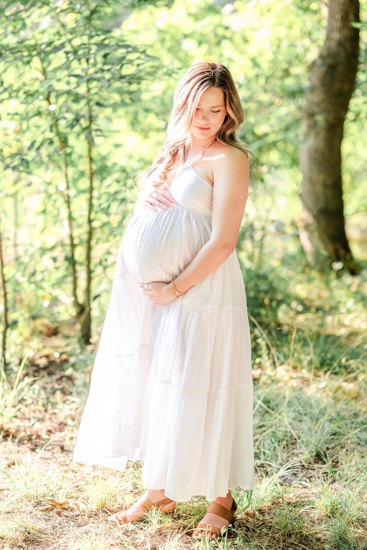 A mama-to-be stands in the sunlight under some trees. She wears a simple white summer dress during her Chesapeake Maternity Photography session.