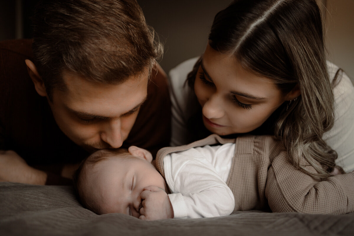 baby ligt op zijn buikje te slapen terwijl papa en mama met hun gezicht boven hun zoontje hangen en liefdevol kijken