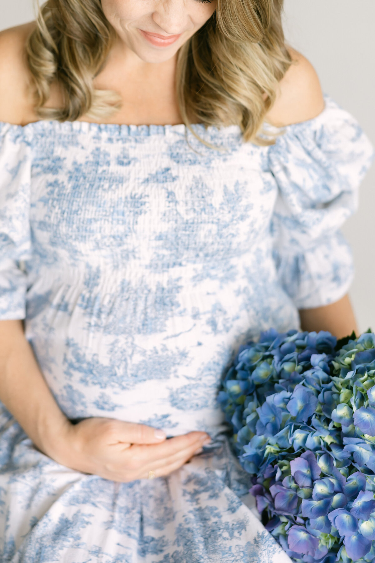 a mother holds her pregnant stomach as she wears a blue and white french toile dress while holding blue hydrangea flowers