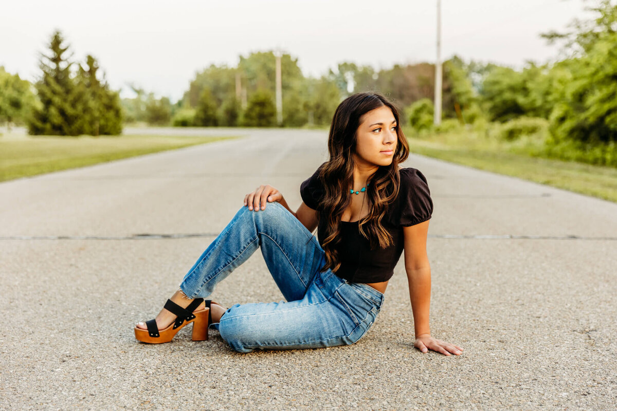 beautiful scene of a teen girl in a black top and jeans with black wedges on sitting in the middle of the road looking over shoulder by Ashley Kalbus Photography