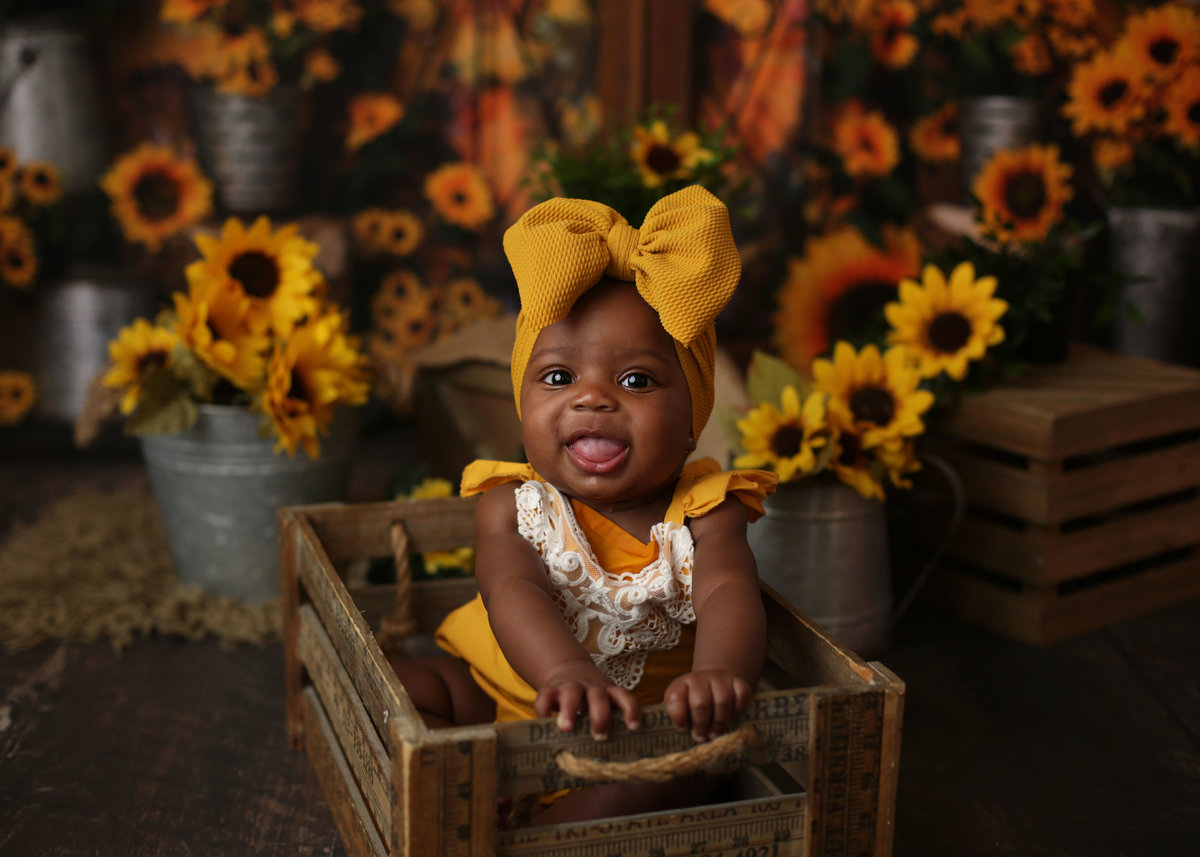 black baby girl sitting in wooden crate with large yellow bow on head sunflowers