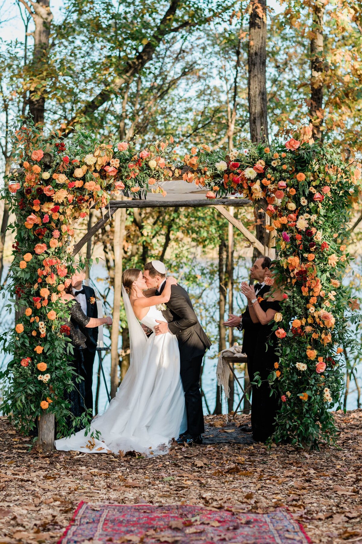 A newlywed couple shares their first kiss as husband and wife under a chuppah adorned with colorful autumnal flowers