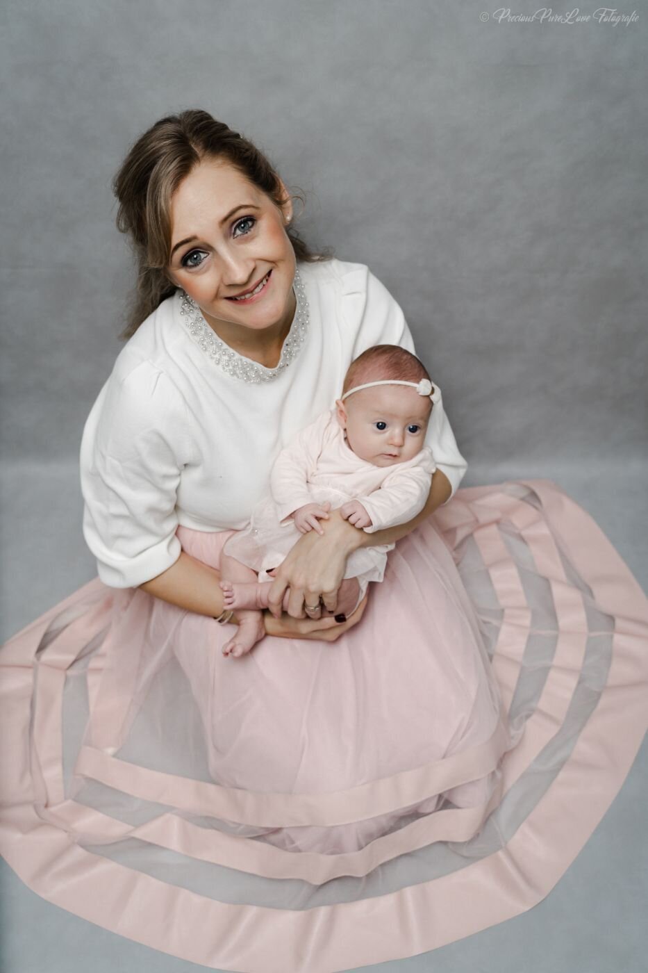 A posed color portrait of a mother sitting on the floor, holding her newborn baby. The mother smiles at the camera, wearing a white top with a beaded collar and a light pink tulle skirt. The baby, in a white outfit and a small bow, looks off to the side. The background is a soft gray.
