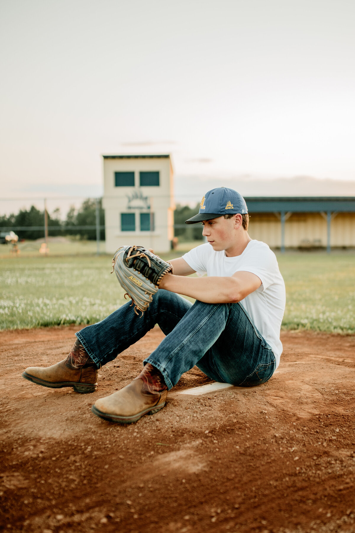 A high school senior sits on a pitcher's mound wearing cowboy boots and jeans during a baseball senior session. Beach Senior