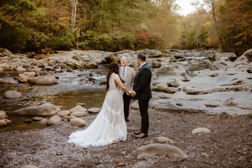 Bride and groom exchanging vows during a riverside ceremony at Greenbrier while eloping to Gatlinburg, standing hand in hand with the officiant amid smooth boulders, flowing water, and vibrant autumn foliage.

