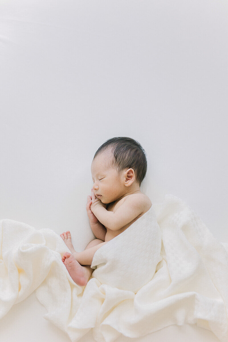 Newborn baby laying on his side with his hands close to his cheeks. He has japanese wraps around him. Taken in Yokohama
