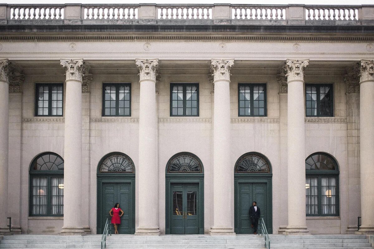 A couple stands apart in front of a classical building with tall columns, each on opposite sides of the grand entrance