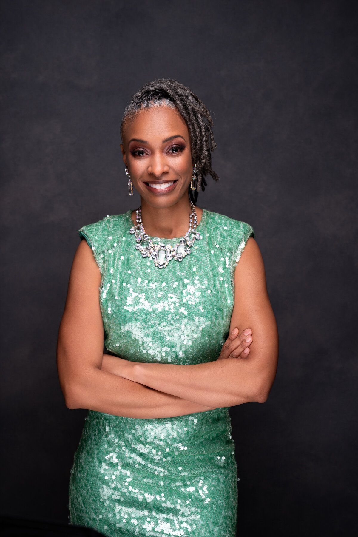 Brown Skinned woman wearing green sequin dress with large diamond necklace while crossing arms and smiling.