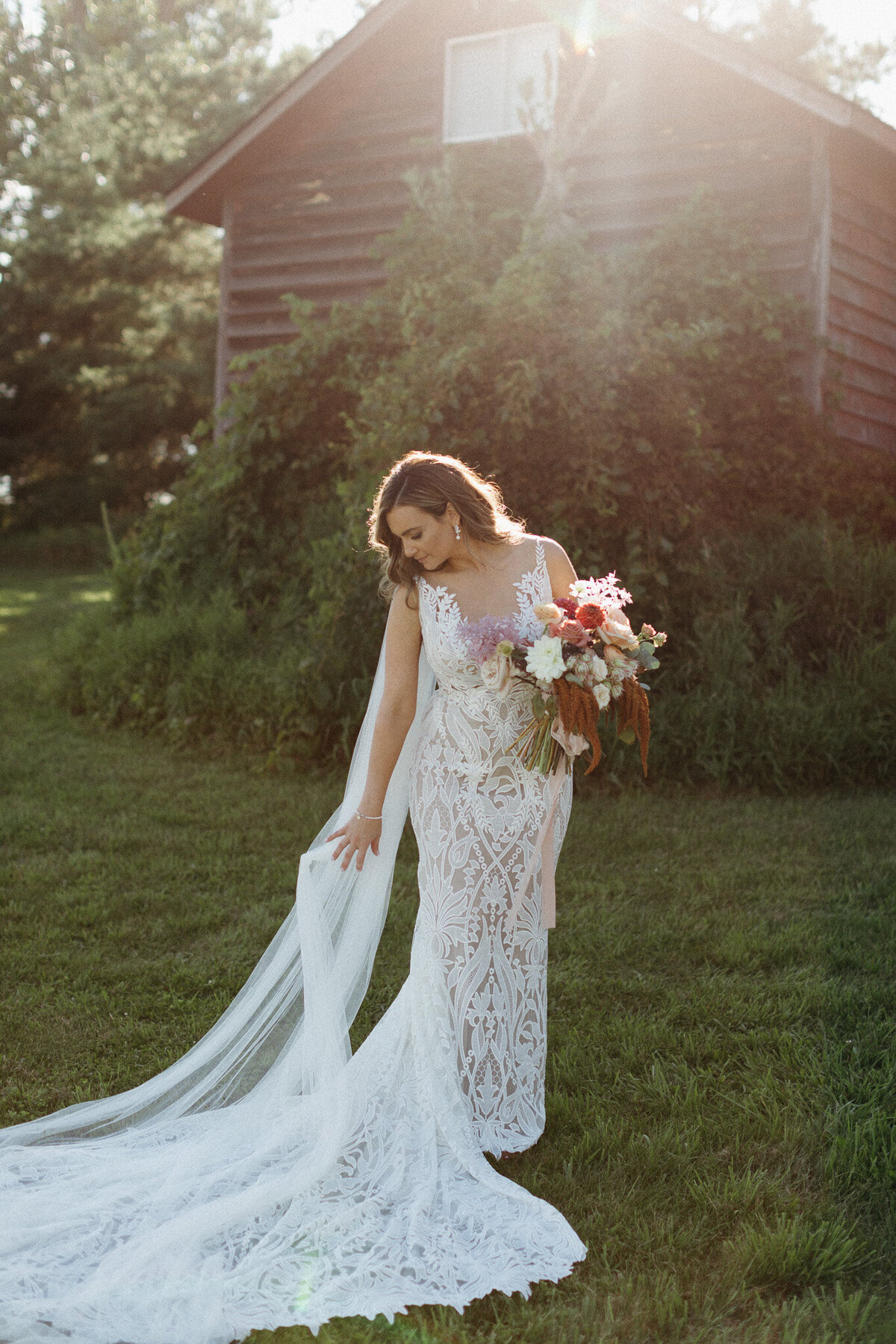 Bride in a detailed lace gown holds a bouquet and gazes down, standing on grass in front of a rustic wooden building with ivy, as sunlight filters through trees—a perfect scene captured by Caledon Valley wedding photographer, Jennifer van Son.