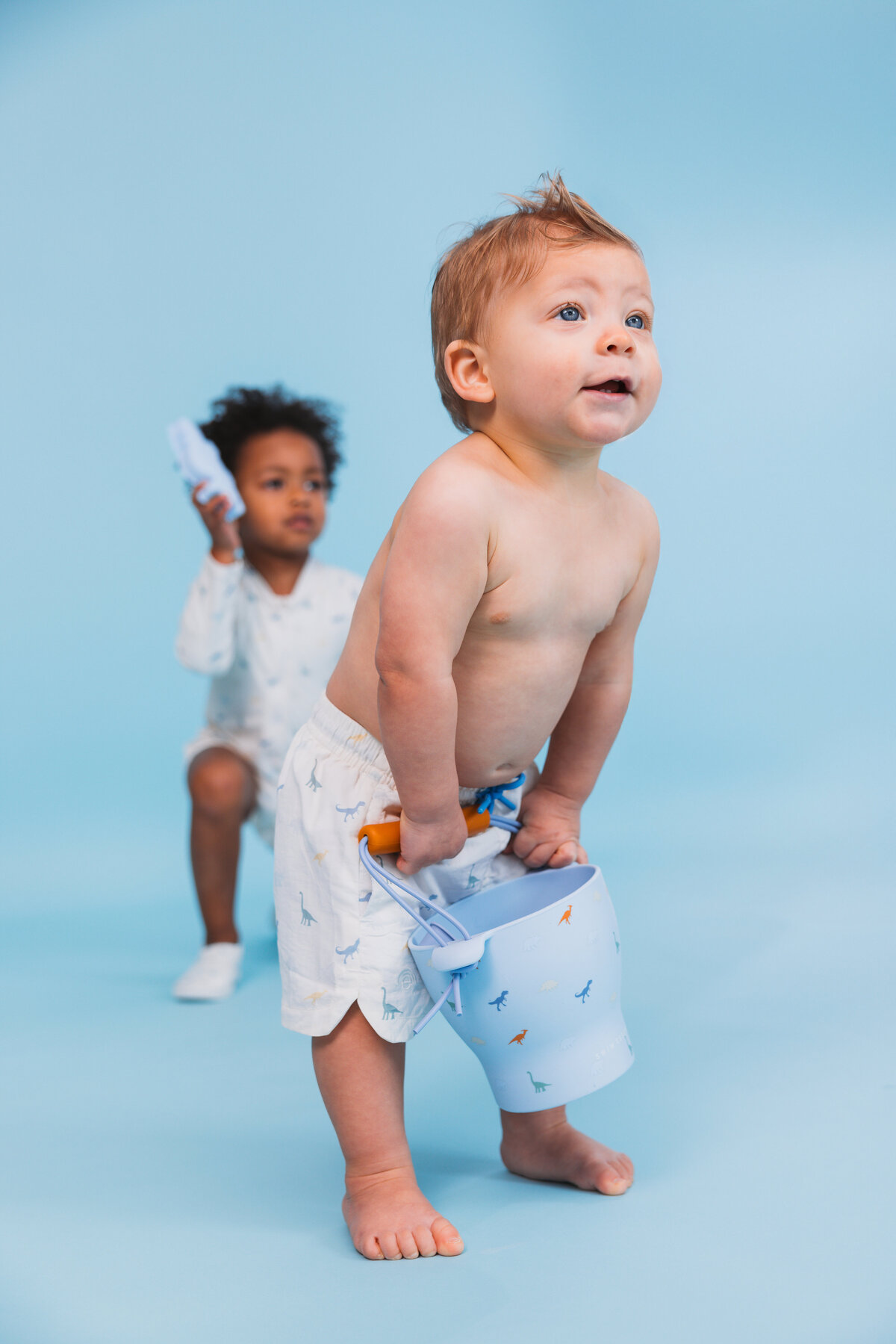 Two young children playing on a blue background, wearing Swim Essentials swimwear with dinosaur prints. One holding a matching blue bucket and the other a toy whale.