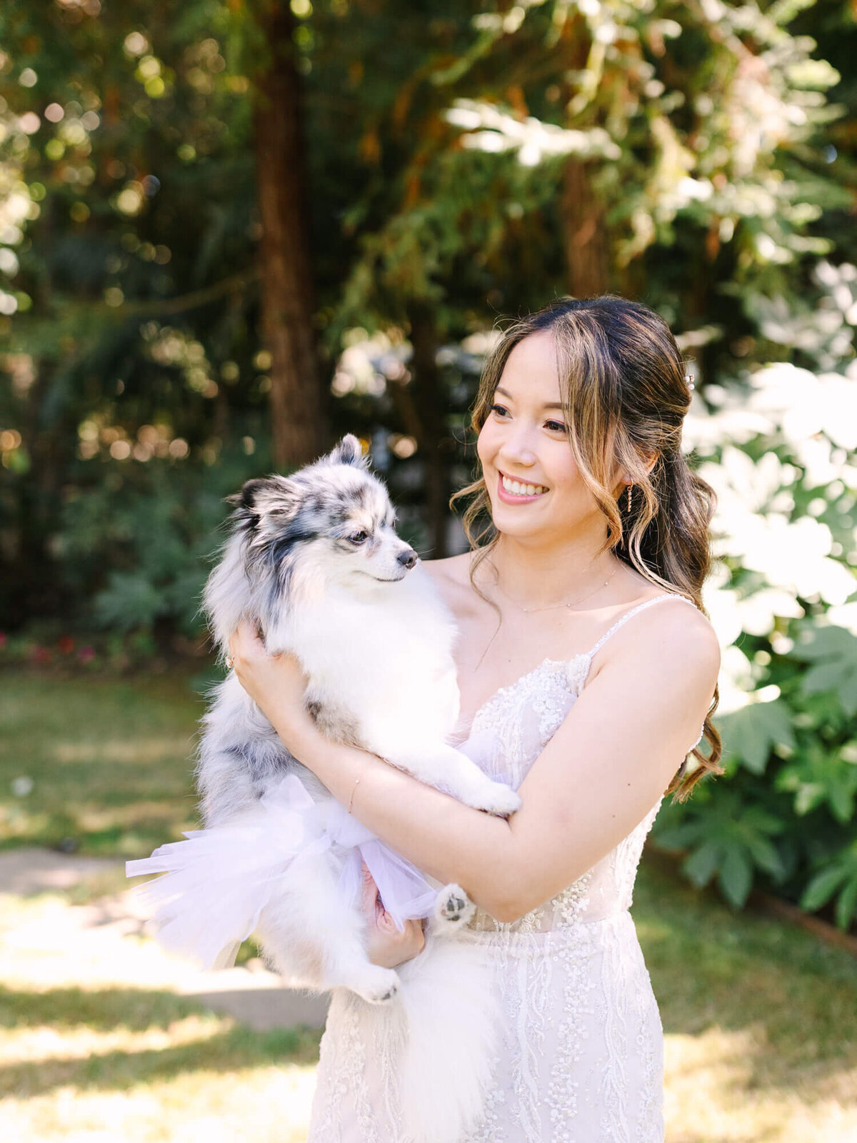A bride in a white lace dress holds a small, fluffy dog in a tutu. They are outdoors with a serene forest backdrop.
