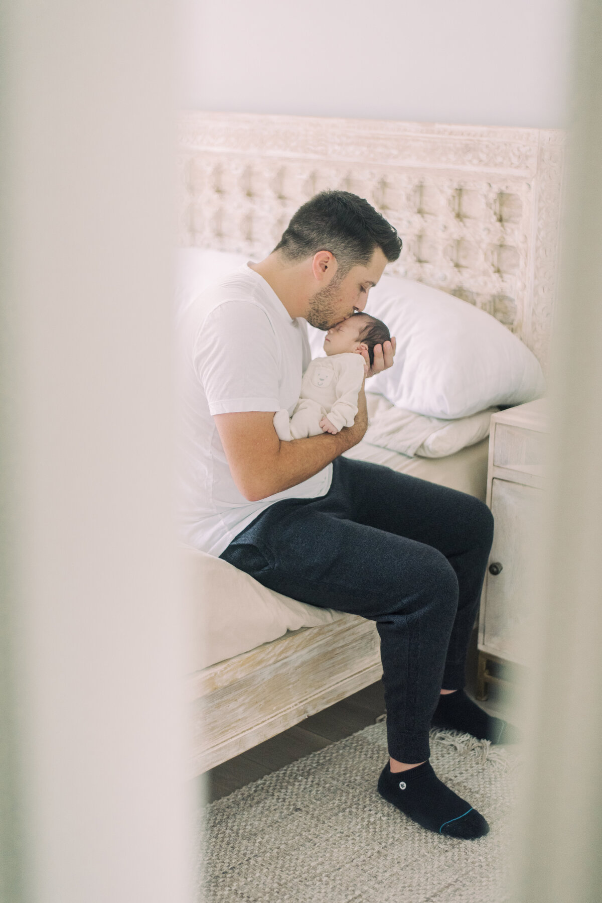 Father kissing his newborn daughter while seated on the bed – peaceful in-home newborn photography in Vaughan