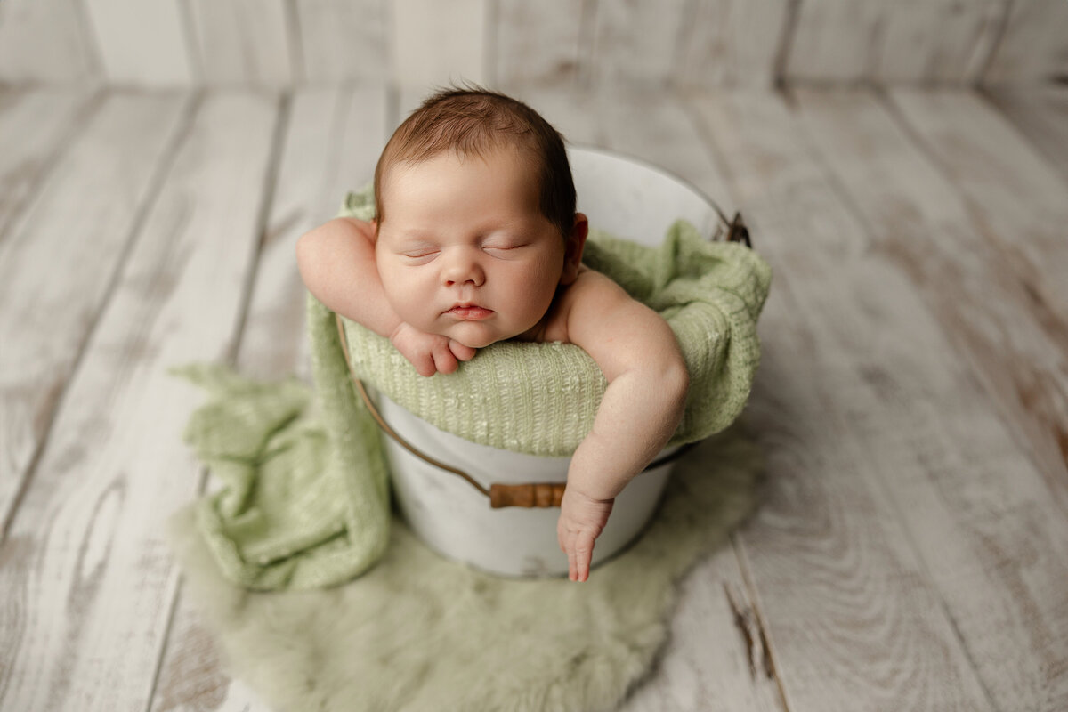 Newborn baby posed with arms draped over a white bucket, surrounded by soft green textures on rustic wood flooring. Captured by Inland Empire newborn photographer Kaitlyn Dawn Photography.