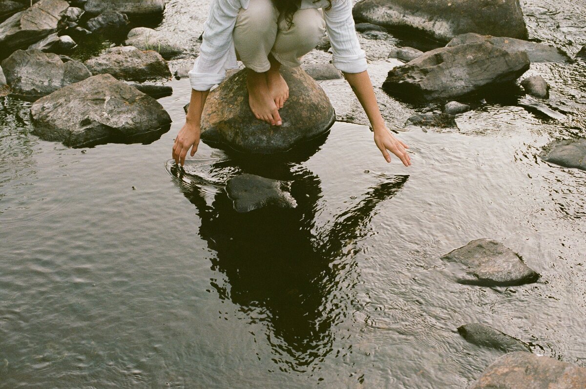 Heather bends down to touch the surface of the river. They are a white person wearing a white outfit.