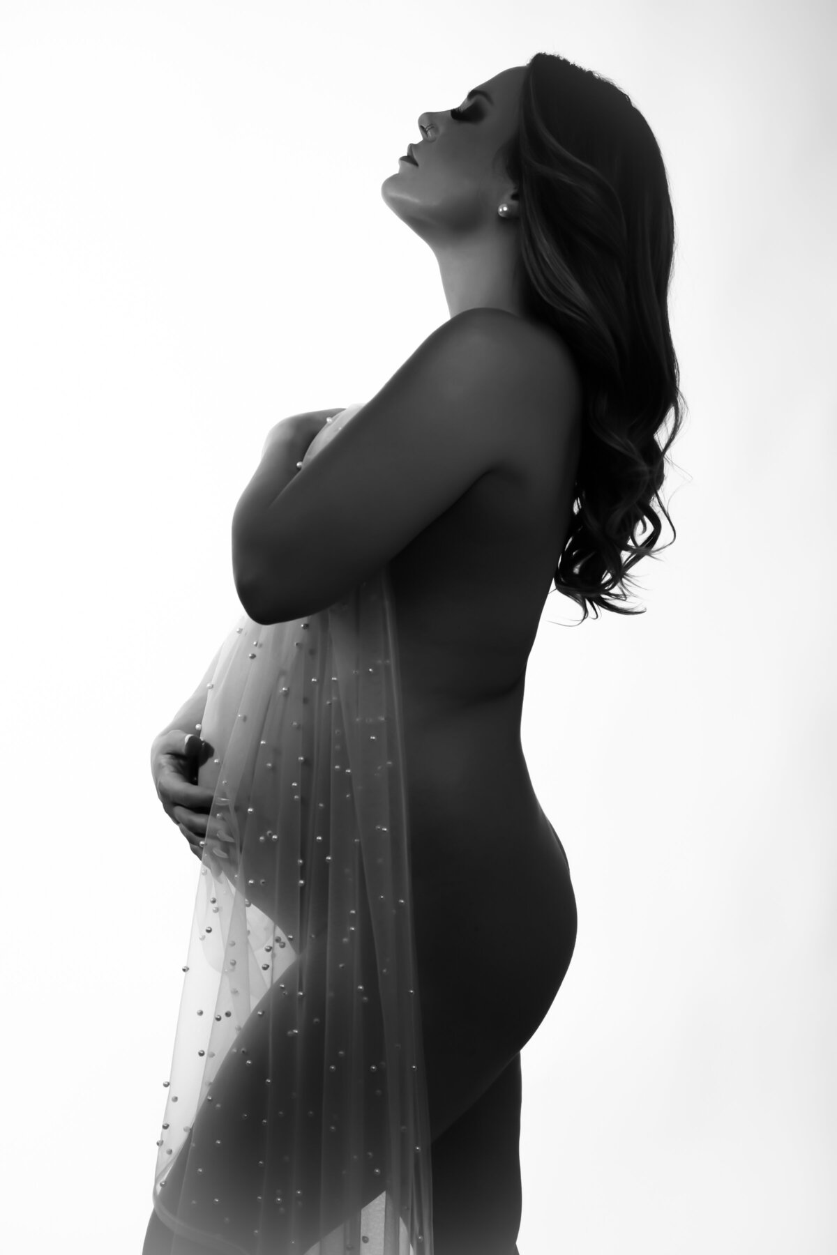 a portrait of a pregnant woman naked wrapped in a sheer pearl fabric touching her belly with one hand and the other covering her breast