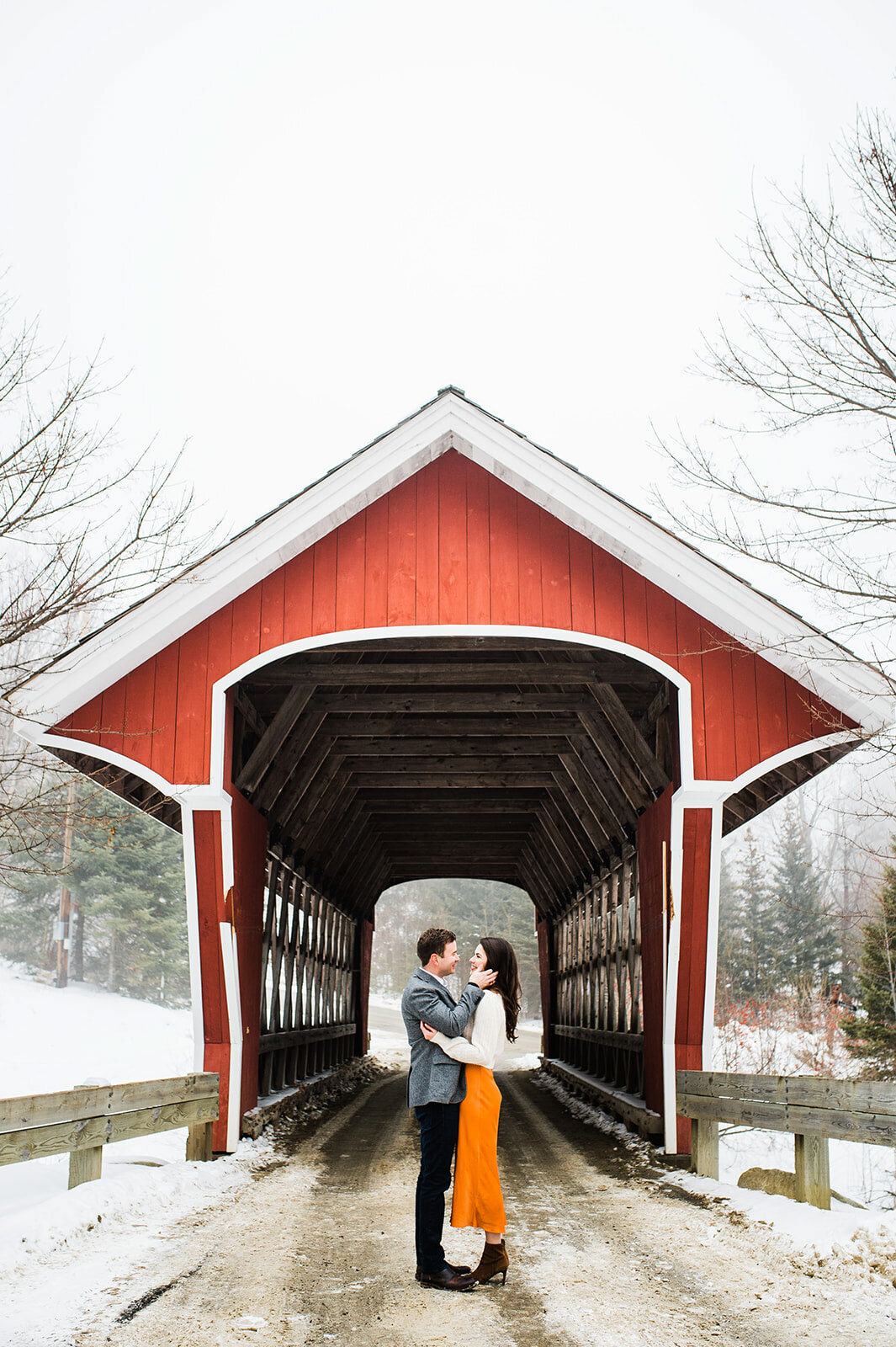 couple kissing under red wooden bridge at stratton mountain vermont in winter