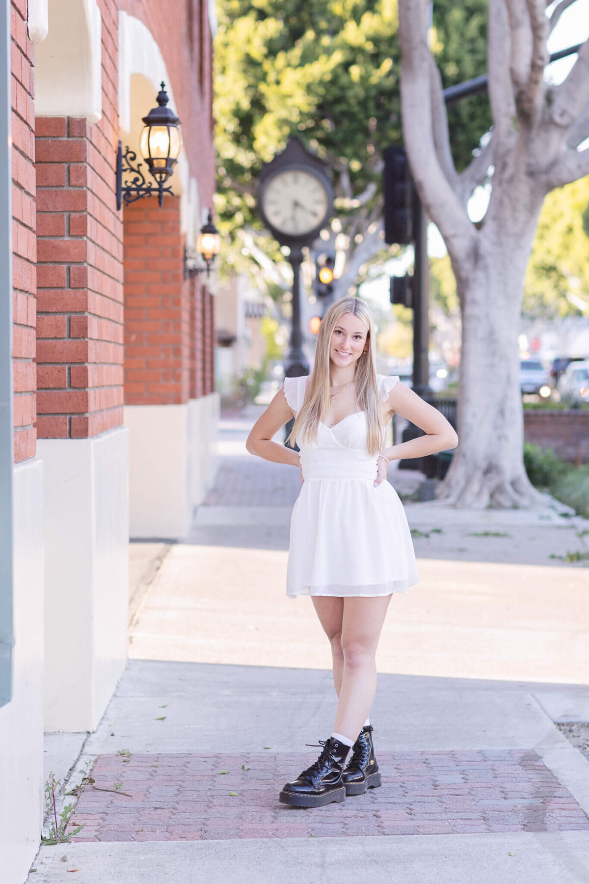 A young woman with long blonde hair stands on a sunlit sidewalk in front of a red brick building, wearing a white dress and black boots. A vintage clock and tree-lined street are in the background.