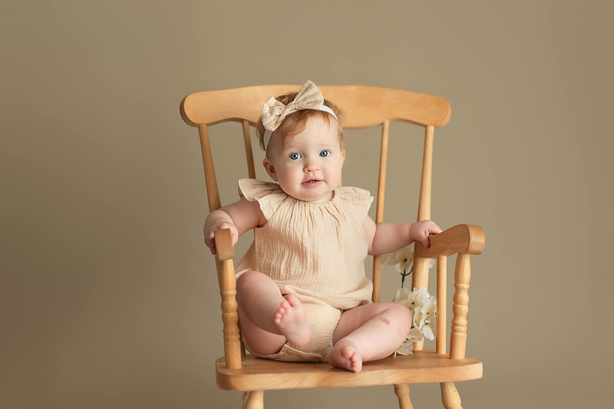 Baby girl dressed in a cream colored romper in a rocking chair.