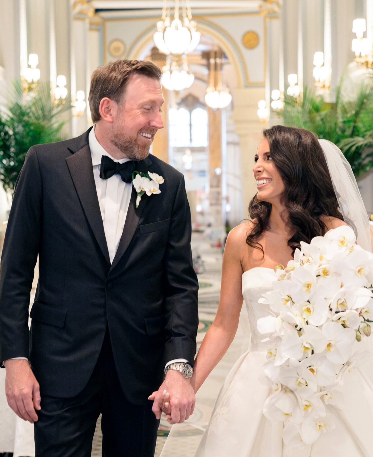 A bride and groom smile at each other while holding hands. The groom wears a black tuxedo with a bow tie, and the bride wears a white gown with a veil, holding a bouquet of white flowers. They stand in an elegant, chandelier-lit venue.