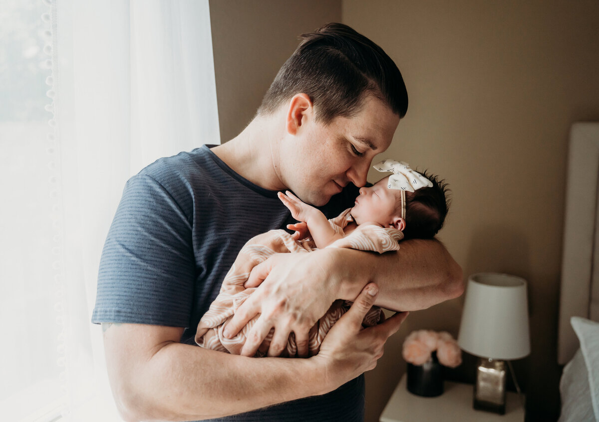 Newborn Photographer, a father stands besides his bed holding his newborn baby daughter
