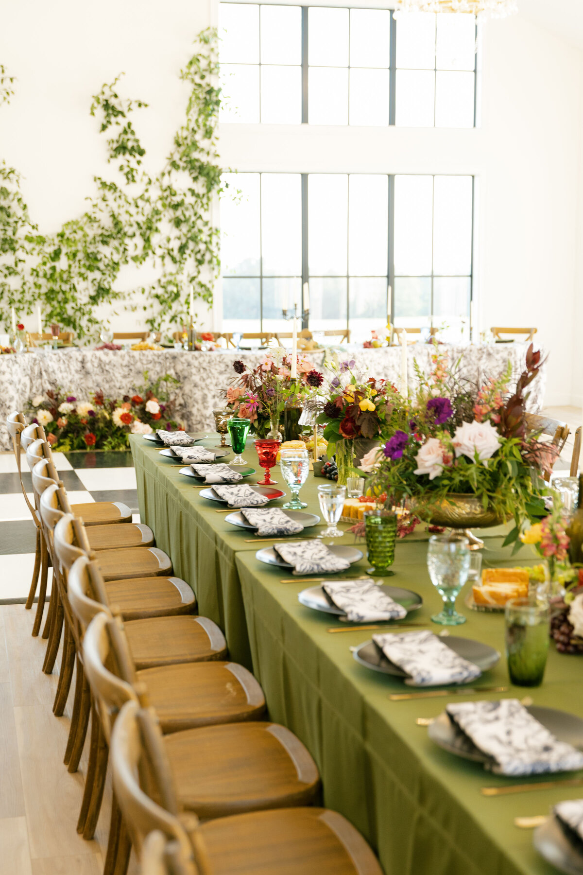 A long wedding reception table decorated with abundant floral arrangements in earthy vessels, featuring roses, greenery, colorful seasonal blooms, taper candles, fruit accents, and curated glassware, styled by a floral designer for an elegant indoor celebration.