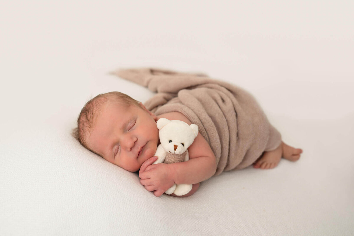 A newborn baby boy wrapped in a brown blanket cuddles his white teddy bear