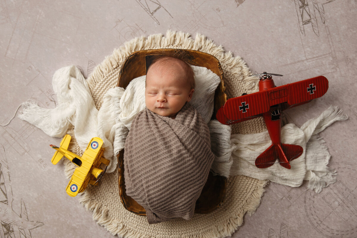 Portrait of a newborn baby wrapped in a blanket and laying in a wooden bowl with toy airplanes next to him