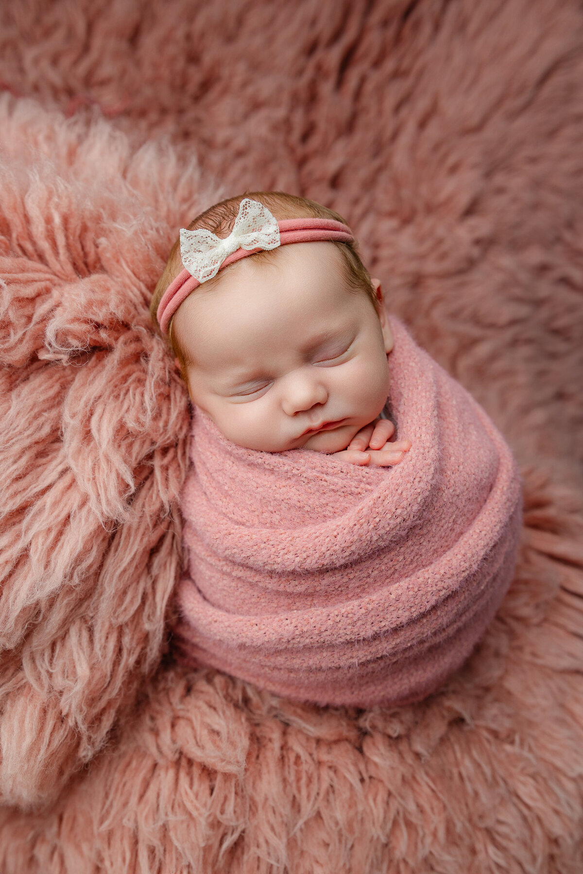 Baby girl swaddled in textured pink fabric, peacefully sleeping on a blush fur layer with a lace bow headband. Photographed in-studio by Inland Empire newborn photographer Kaitlyn Dawn.