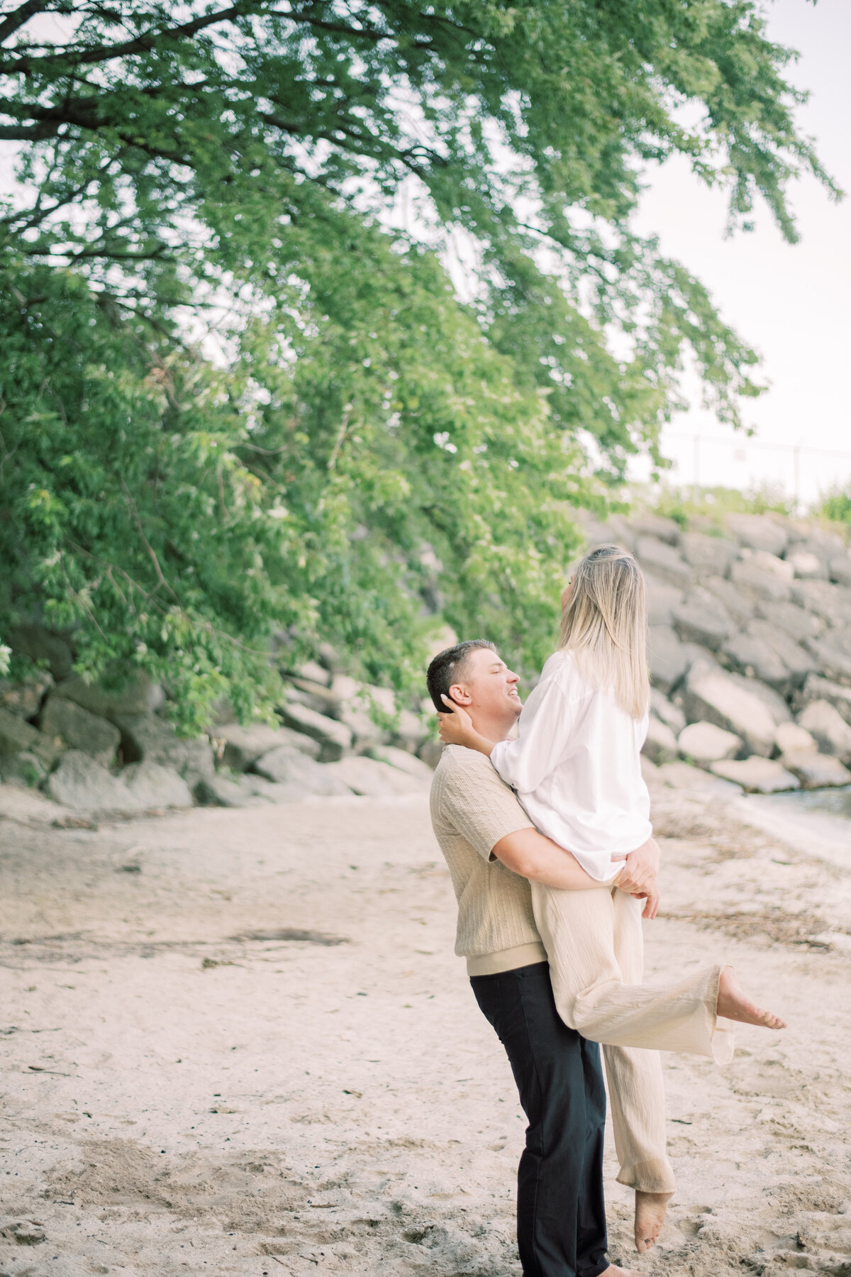 Engaged couple with their golden doodle by the lakeshore in Oakville, all smiles as they enjoy a quiet moment together