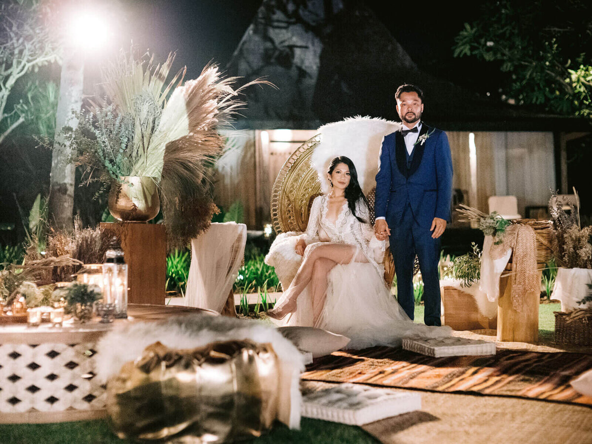 The bride is seated in a native wooden chair, the groom is standing beside her in Khayangan Estate, Bali, Indonesia. Image by Jenny Fu Studio