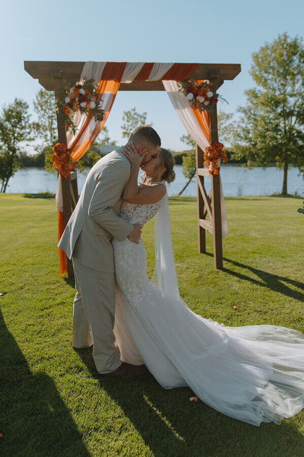 A bride and groom kiss during their outdoor wedding ceremony in front of a wooden arch decorated with orange fabrics and florals.